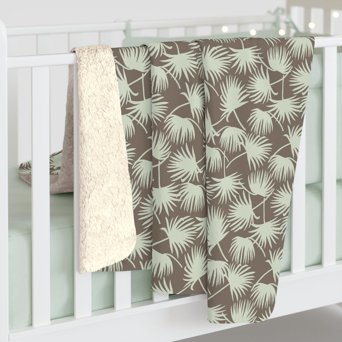 Fan Palm Mocha Mint sherpa fleece blanket with a repeating mint palm leaf pattern on a warm mocha background hanging off a white crib.