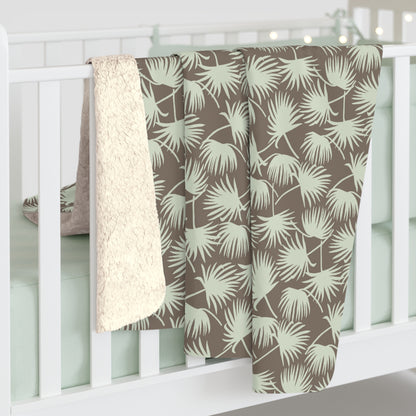 Fan Palm Mocha Mint sherpa fleece blanket with a repeating mint palm leaf pattern on a warm mocha background hanging off a white crib.