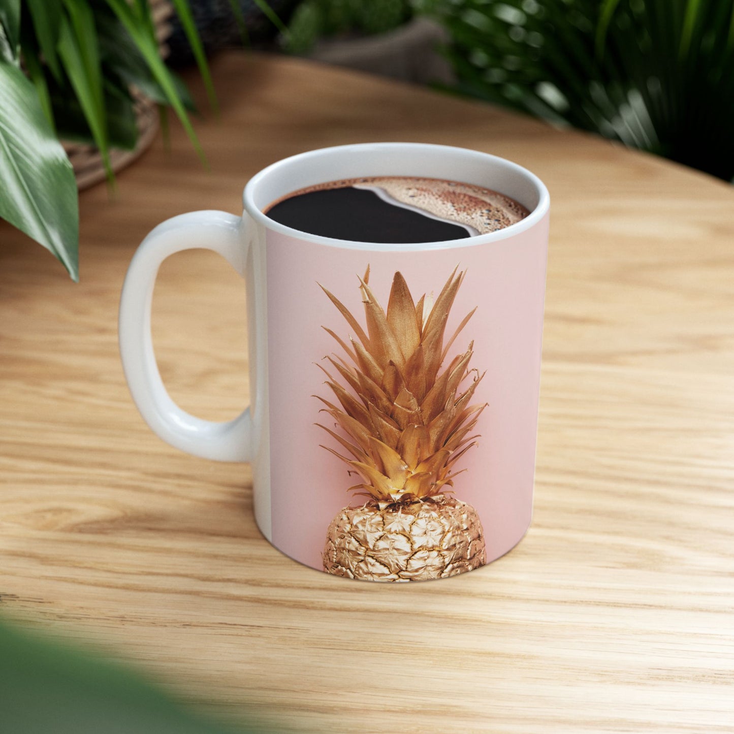 A white ceramic coffee mug filled with black coffee, featuring a printed design of a golden pineapple on a dusty pink background, set on a light wood table with green plants in the background.