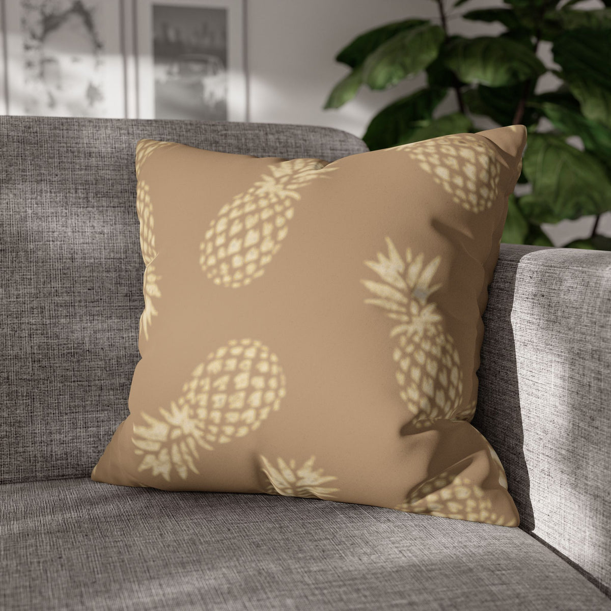 A square throw pillow with a beige and cream pineapple pattern, resting on a gray textured sofa with a green plant in the background.