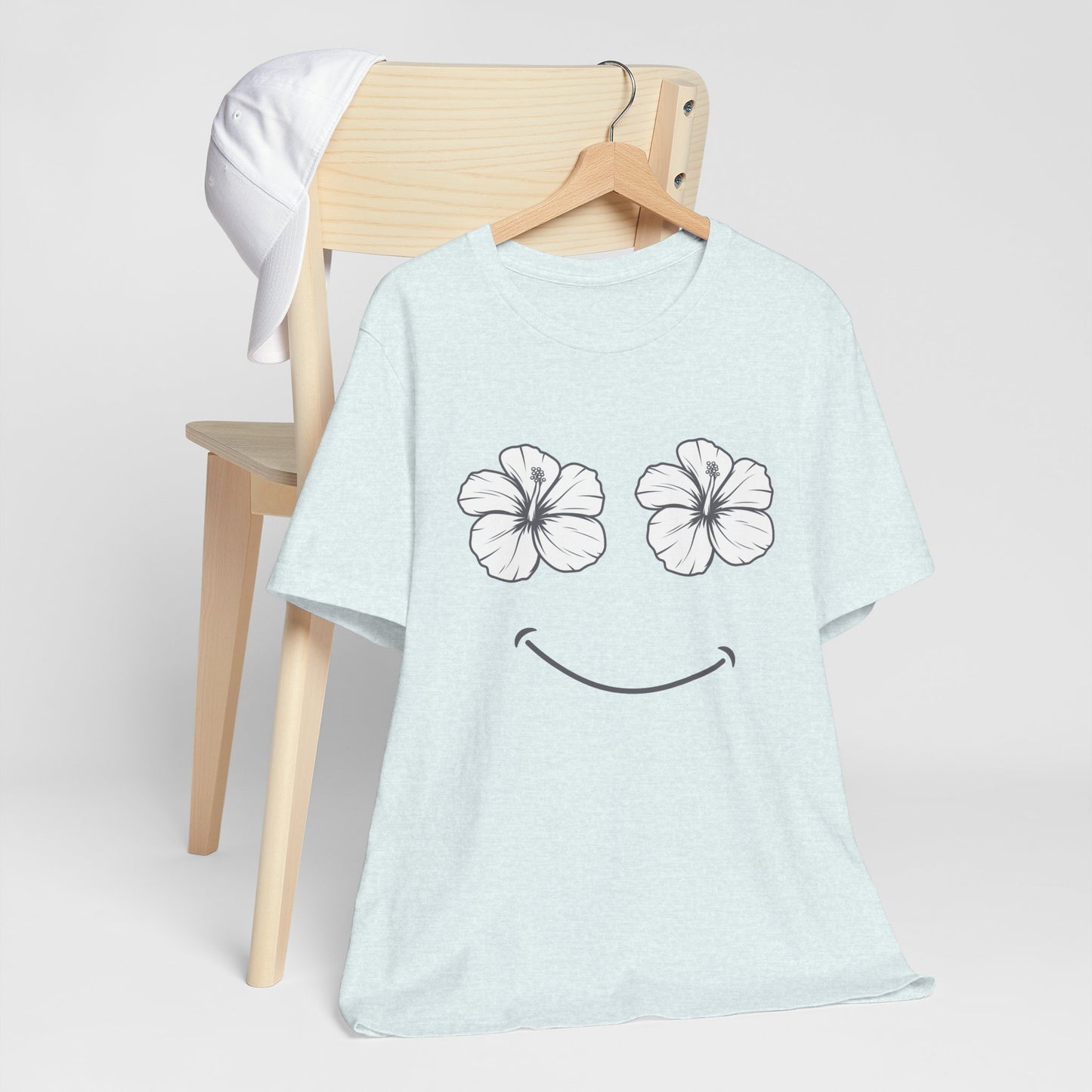 A light blue heather t-shirt with a black smiley face graphic made of two hibiscus flowers for eyes, hanging on a wooden hanger on a light wood chair.