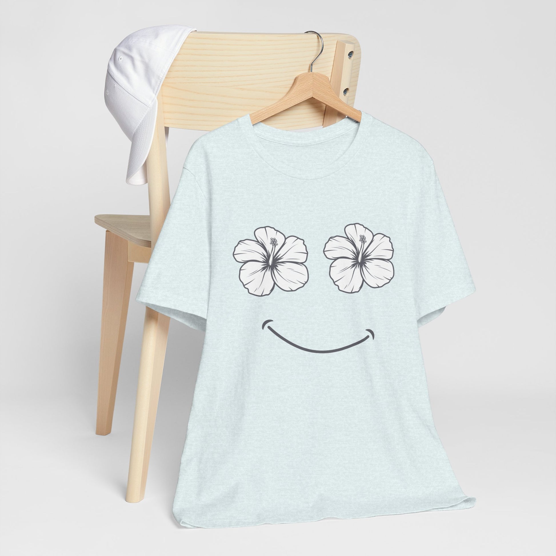 A light blue heather t-shirt with a black smiley face graphic made of two hibiscus flowers for eyes, hanging on a wooden hanger on a light wood chair.