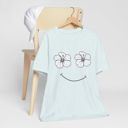 A light blue heather t-shirt with a black smiley face graphic made of two hibiscus flowers for eyes, hanging on a wooden hanger on a light wood chair.