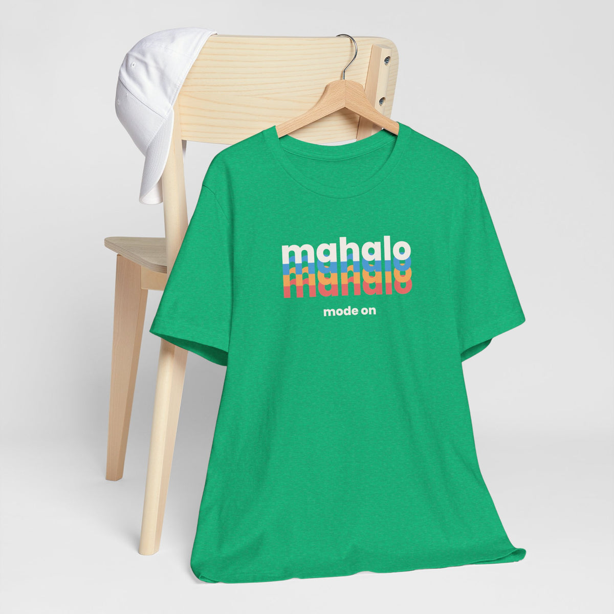 A bright green short-sleeve t-shirt on a wooden hanger, styled in front of a light wood chair with a white cap draped over the backrest. The shirt features a retro-style graphic on the chest reading 'mahalo mode' in white and rainbow stripes, with 'mode on' printed below.