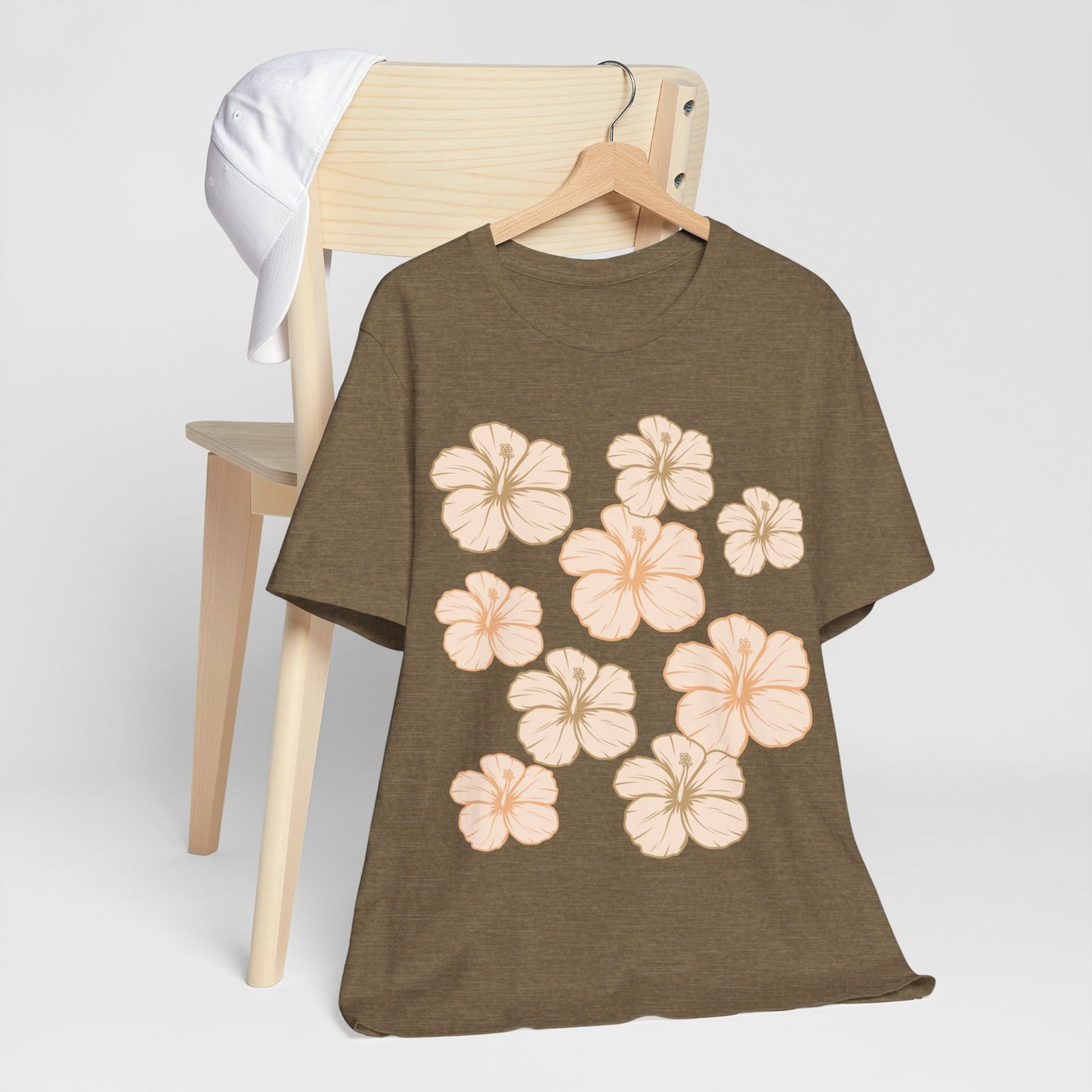 Olive Hibiscus All Over Graphic Tee featuring scattered tropical hibiscus flowers on a soft unisex tee inspired by island life in Hawaii hanging on a wood chair.