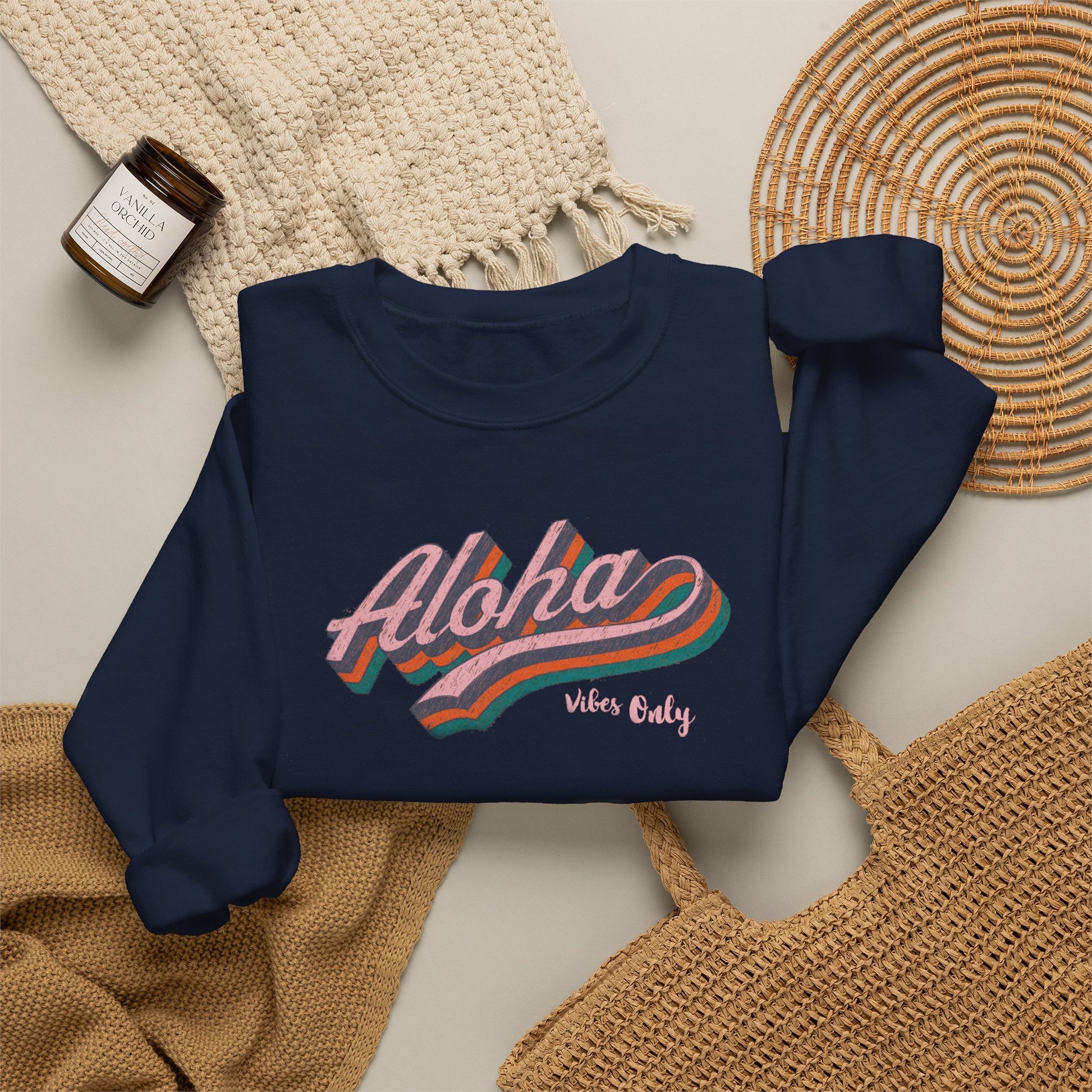 A navy blue sweatshirt with a retro-style 'Aloha Vibe's Only' graphic in pink, orange, and teal, styled in a flat lay with a beige knit blanket, a woven placemat, and a small candle.
