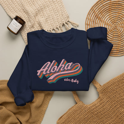 A navy blue sweatshirt with a retro-style 'Aloha Vibe's Only' graphic in pink, orange, and teal, styled in a flat lay with a beige knit blanket, a woven placemat, and a small candle.