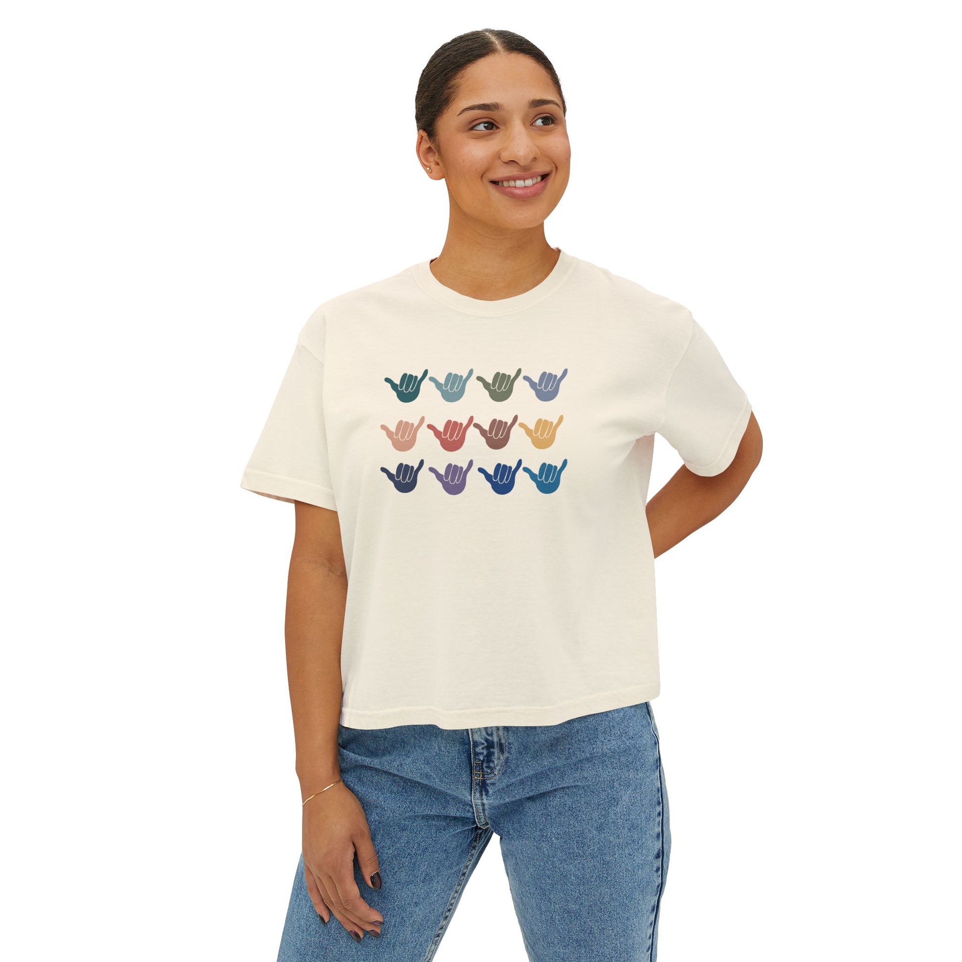 A woman wearing an off-white crop short-sleeve t-shirt with a graphic of twelve shaka signs in various colors arranged in three rows, displayed on a clean white background.
