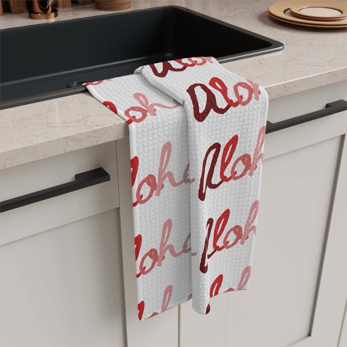 Red Aloha Hawaiʻi kitchen towel with repeating script design in shades of red on white hanging off a kitchen sink