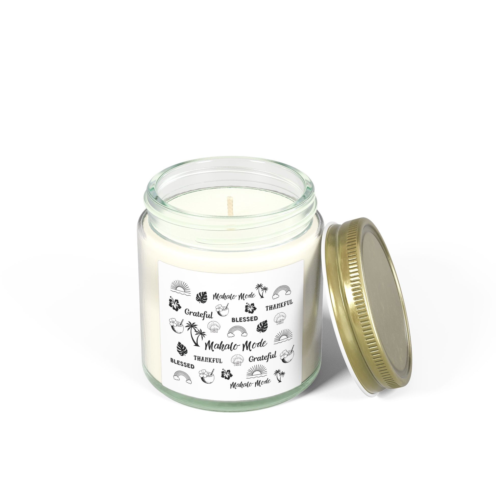 A glass jar candle with a gold metal lid and a white paper label featuring black tropical-themed illustrations and text like 'Mahalo Mode,' 'Grateful,' and 'Blessed,' shown on a clean white background.
