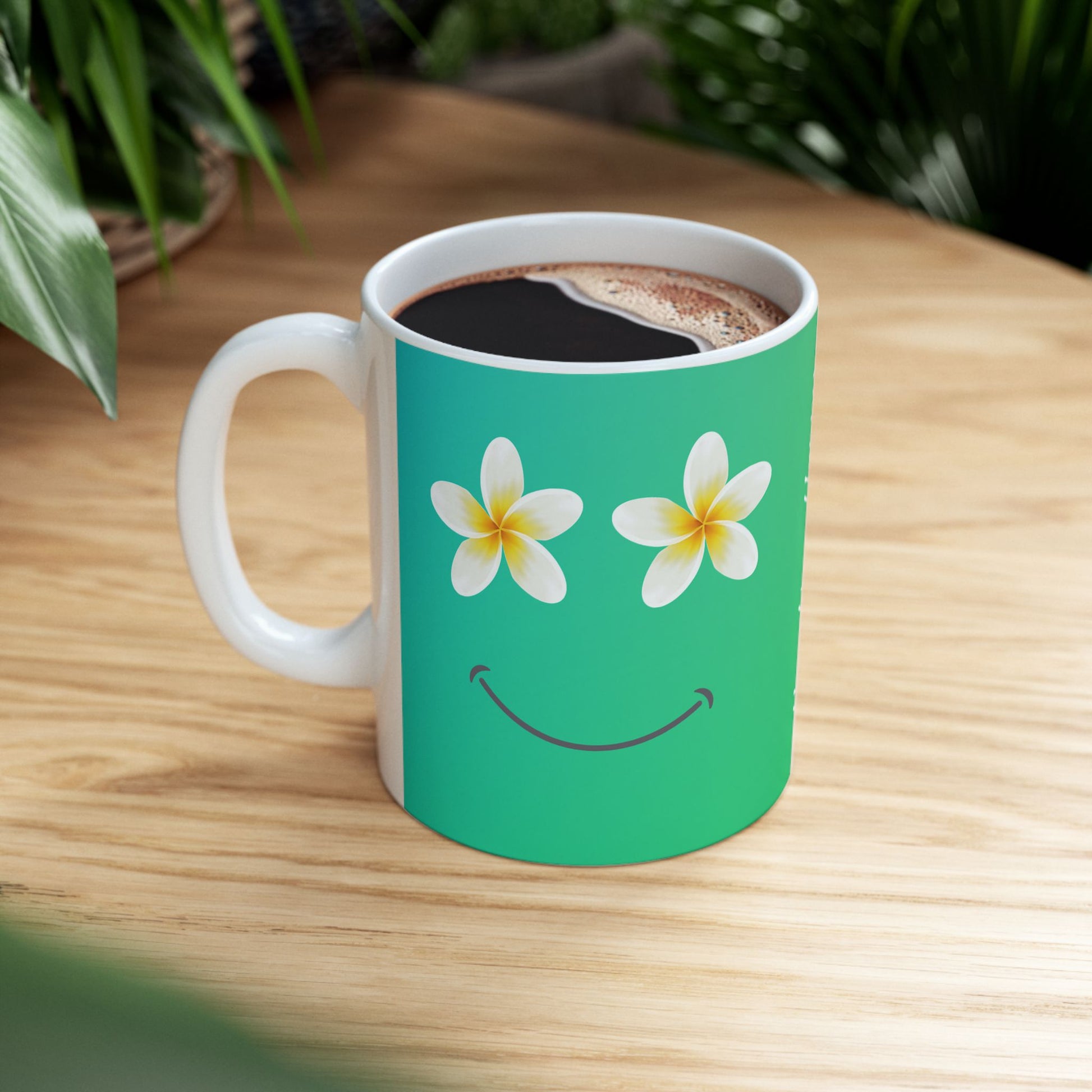 A white ceramic coffee mug with a bright green front panel featuring a smiling face made of two white plumeria flowers with yellow centers. The mug is filled with black coffee and sits on a light wood table with blurred green plants in the background.