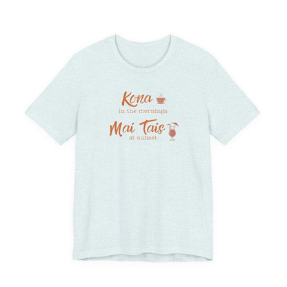 A light heather blue short-sleeve t-shirt with a graphic print on the front reading 'Kona in the mornings Mai Tais at sunset' in orange script, accompanied by small illustrations of a coffee cup and a cocktail glass.