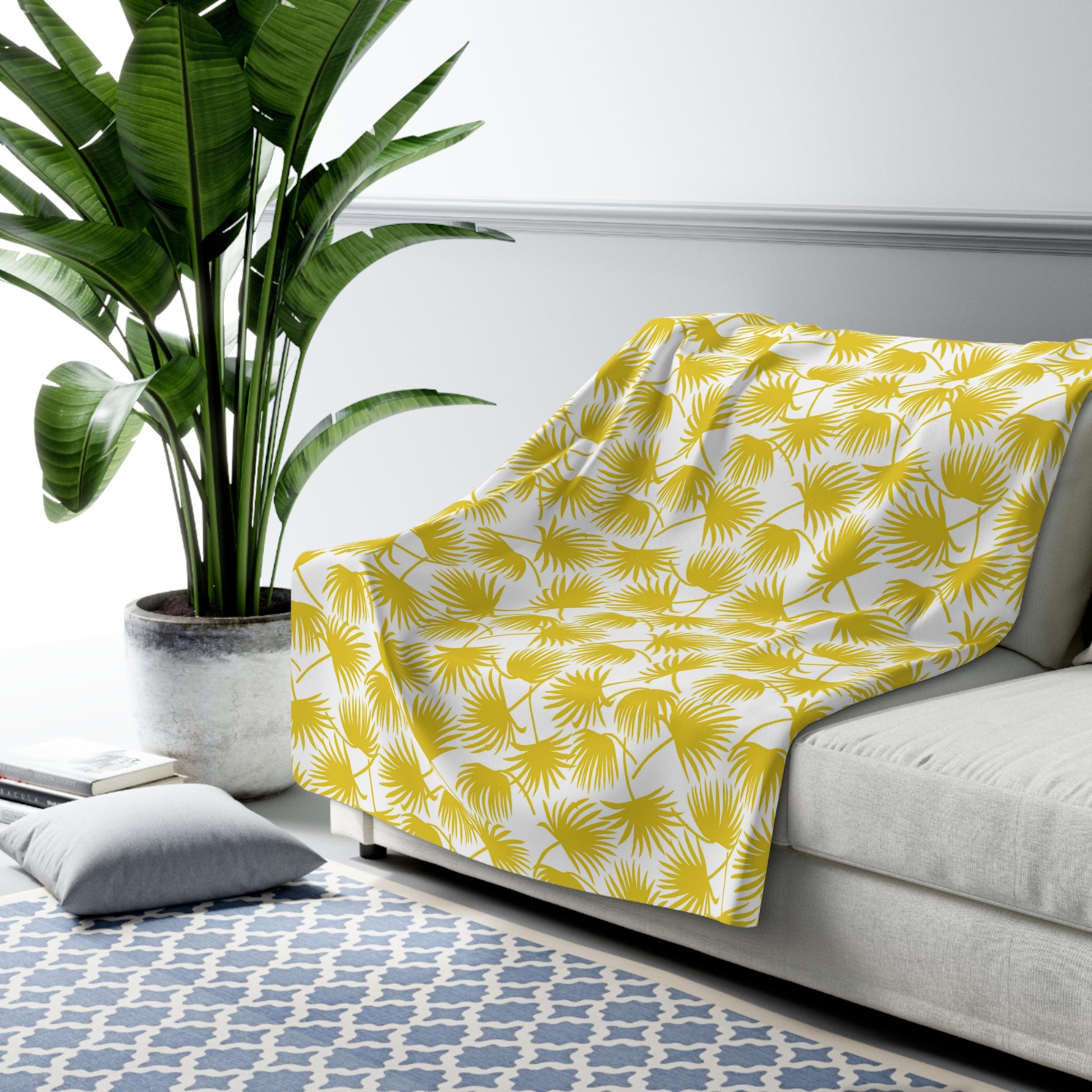A yellow and white palm leaf patterned throw blanket is draped over a light gray sofa in a modern living room setting with a potted plant, blue patterned rug, and gray pillow.