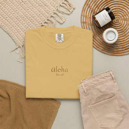A folded pale yellow t-shirt with 'Aloha for all' printed on the chest, styled in a flat lay with beige pants, a woven placemat, a candle, and a knit blanket.