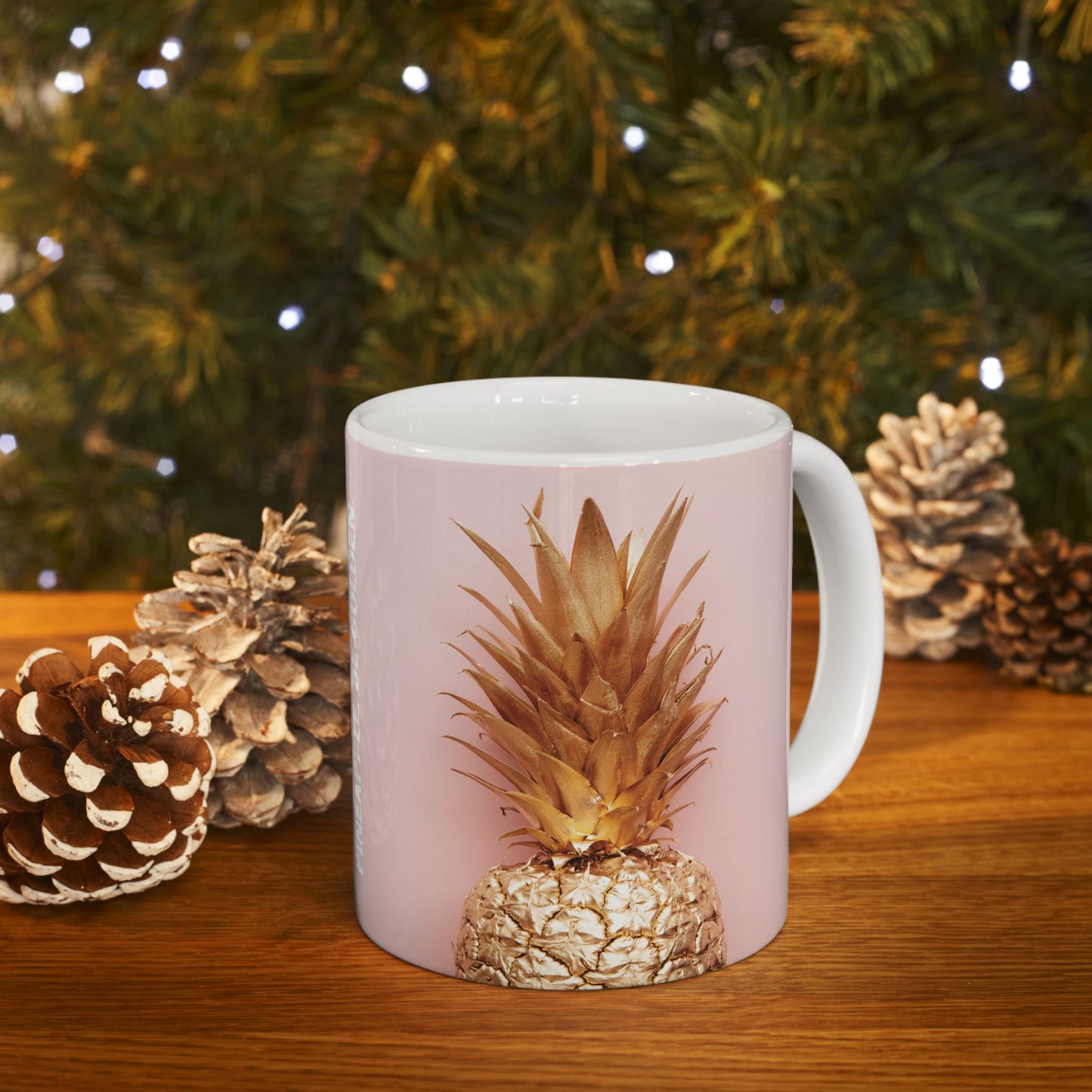 A white ceramic mug with a light pink printed wrap, featuring a graphic of a pineapple with metallic gold glitter details, set on a wooden table with pinecones and a blurred Christmas tree background.