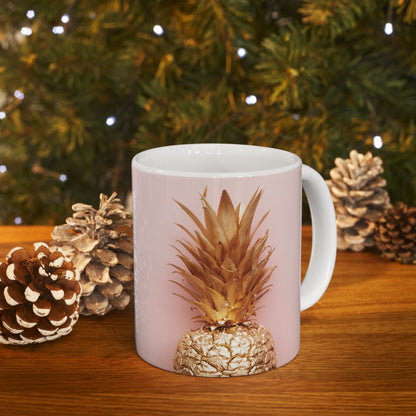 A white ceramic mug with a light pink printed wrap, featuring a graphic of a pineapple with metallic gold glitter details, set on a wooden table with pinecones and a blurred Christmas tree background.