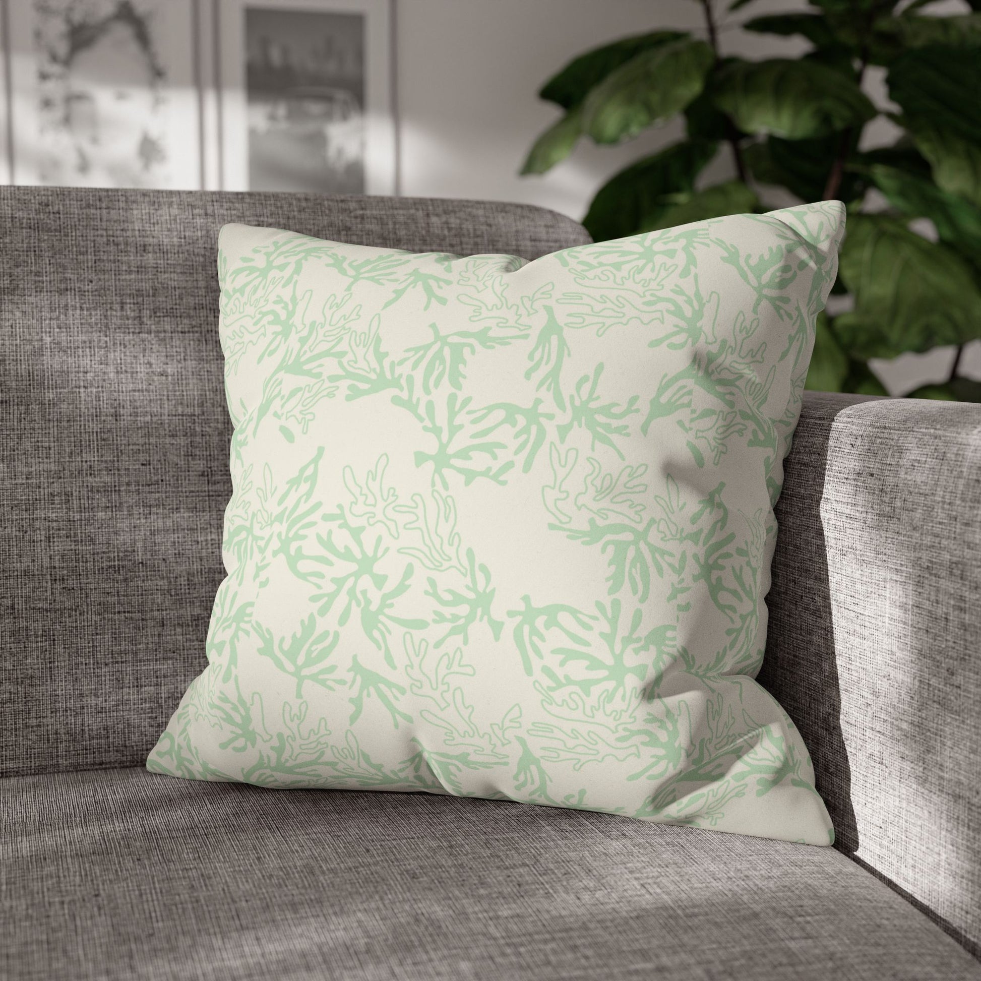 A square throw pillow with a light sage green coral pattern on an off-white background, resting on a textured gray fabric sofa with a green houseplant in the background.