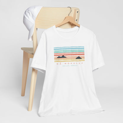A hanging white t-shirt with a colorful retro sunset graphic and the text 'NÁ MOKULUA KAILUA, HAWAII' on the front, hanging from a wood chair.