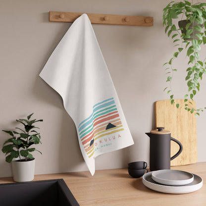 A white kitchen towel with a colorful retro sunset stripe pattern and the text 'NA MOKULUA HAWAII' hanging on a wooden wall rack above a kitchen counter with dishes and plants.