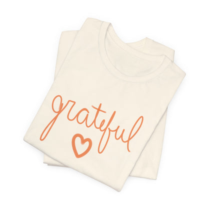 A folded cream-colored t-shirt with the word 'grateful' written in orange cursive script and a matching orange heart outline underneath, displayed on a clean white background.