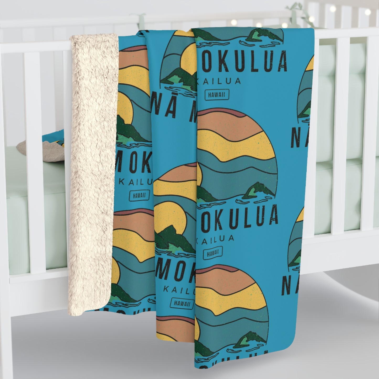 A baby blanket draped over a white crib, featuring a blue background with a retro sunset illustration and the words 'MOKULUA KAILUA HAWAII'. The blanket has a soft, textured cream sherpa backing visible on the left side.