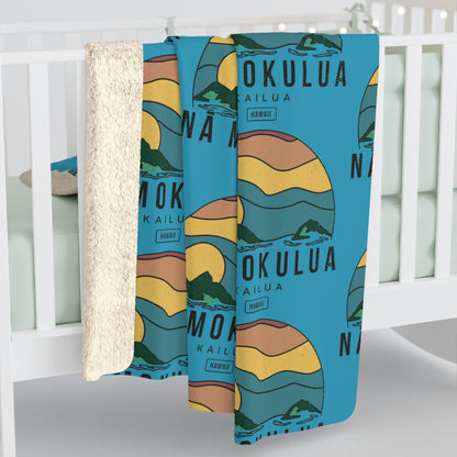 A baby blanket draped over a white crib, featuring a blue background with a retro sunset illustration and the words 'MOKULUA KAILUA HAWAII'. The blanket has a soft, textured cream sherpa backing visible on the left side.