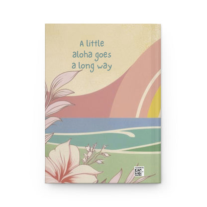 A hardcover notebook with a retro-style tropical illustration on the cover, featuring the text 'A little aloha goes a long way' in blue. The cover has a warm beige background with pink, blue, green, and yellow abstract landscape shapes and floral illustrations.