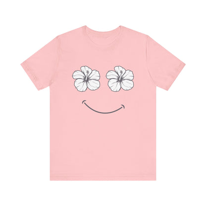 A light pink short-sleeve t-shirt with a graphic of two white hibiscus flowers forming eyes and a simple black smiley mouth on the front, displayed against a solid white background.