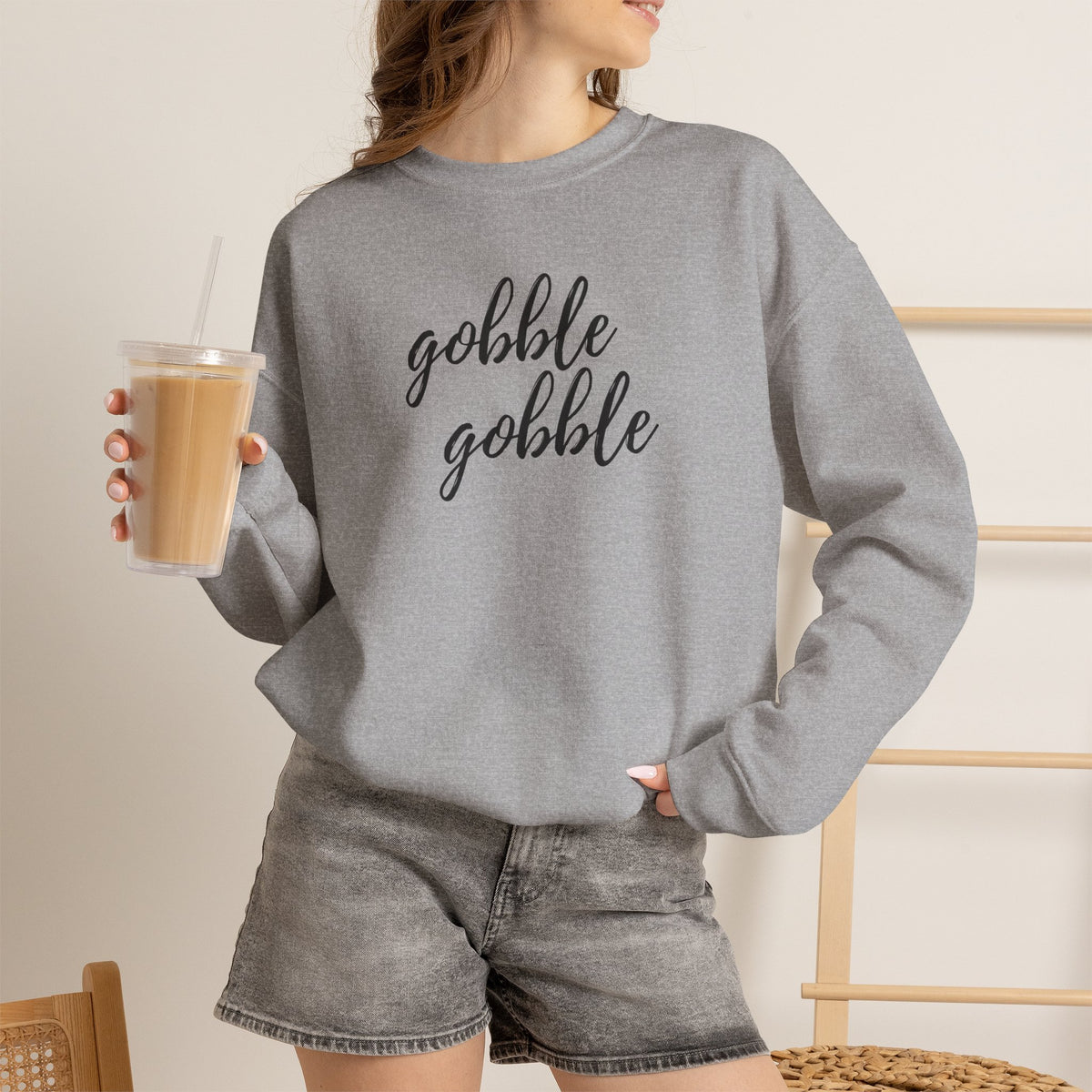 A woman wearing a sport grey crewneck sweatshirt with a graphic that reads 'Gobble Gobble' in black. She is holding a iced coffee and wearing gray denim shorts in a brightly lit indoor setting.