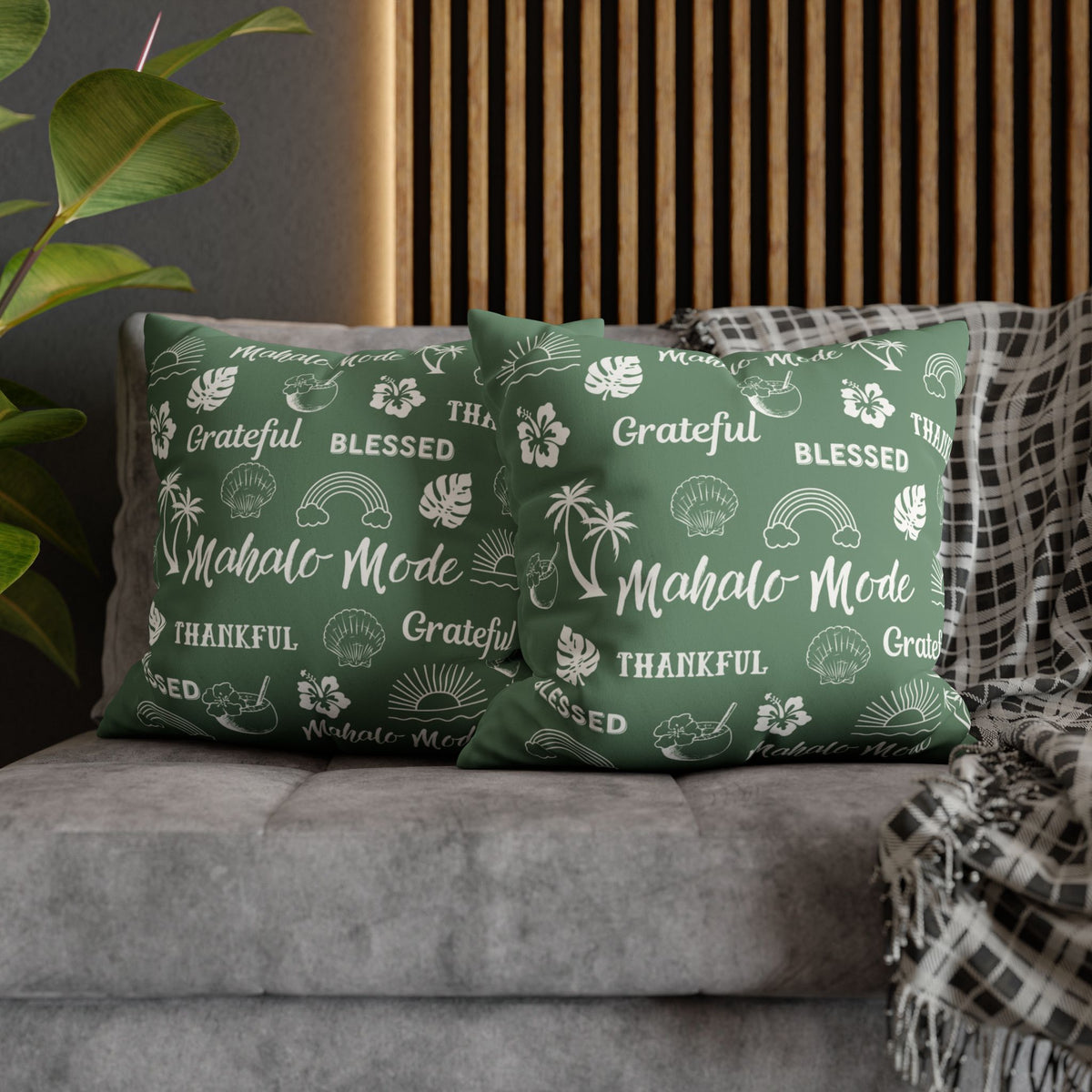Two green throw pillows with white Hawaiian-themed text and illustrations, styled on a gray sofa with a wooden slat wall and a plaid throw blanket in the background.