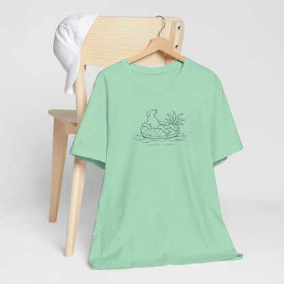 Mint green t-shirt with a graphic design of a dog on a pineapple floatie with text ʻKAILUA HAWAIIʻ on a wooden chair against a white background