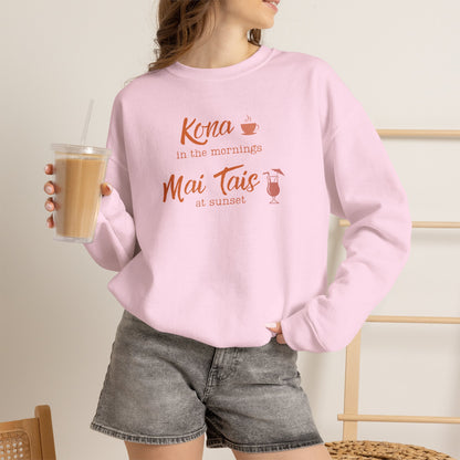 A woman holding an iced coffee wearing a light pink crewneck sweatshirt featuring orange text that reads 'Kona in the mornings Mai Tais at sunset' with small coffee cup and cocktail glass illustrations.