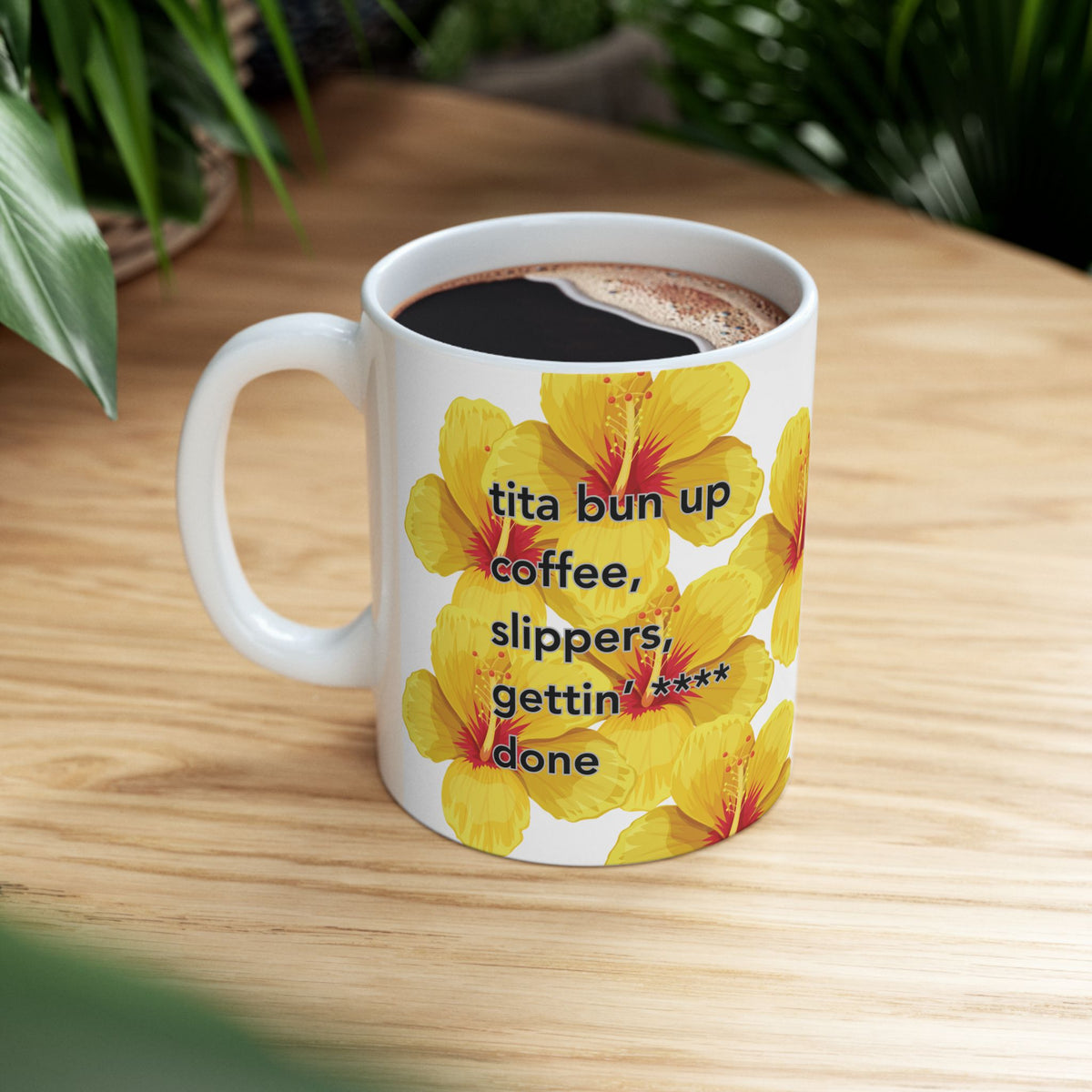 Tita Bun coffee mug with yellow hibiscus design by Global Village Kailua sitting on wood table with green plants.
