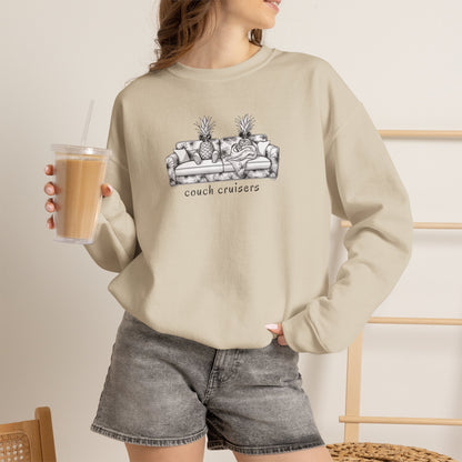 Woman wearing a sand crewneck sweatshirt featuring a black and white line art graphic of a couch with pineapples and the text 'couch cruisers' on the chest. She is holding an iced coffee.