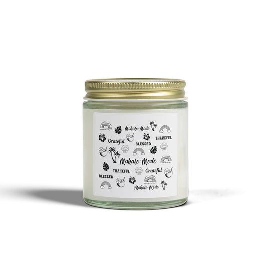 A glass jar candle with a gold metal lid and a white paper label featuring black tropical-themed illustrations and text like 'Mahalo Mode,' 'Grateful,' and 'Blessed,' shown on a clean white background.