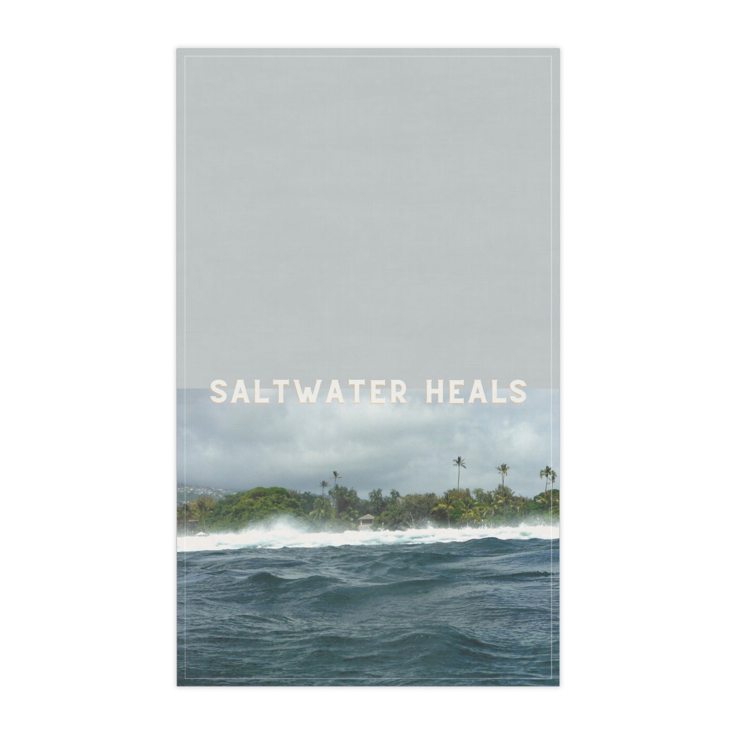 A vertical tea towelfeaturing a seascape with ocean waves, a distant tropical shoreline with palm trees, and the text 'SALTWATER HEALS' across the center. The top portion is a muted light gray, while the lower portion shows blue-gray water and green-brown land under a cloudy sky.