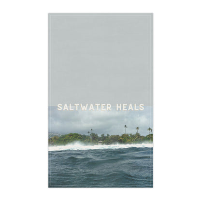A vertical tea towelfeaturing a seascape with ocean waves, a distant tropical shoreline with palm trees, and the text 'SALTWATER HEALS' across the center. The top portion is a muted light gray, while the lower portion shows blue-gray water and green-brown land under a cloudy sky.