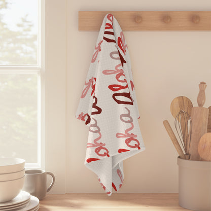 Red Aloha Hawaiʻi kitchen towel with repeating script design in shades of red on white hanging off a wooden peg in bright kitchen