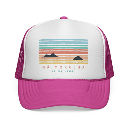 A pink and white trucker hat featuring a colorful graphic of a sunset over mountains with the text 'NÁ MOKULUA KAILUA, HAWAII'.