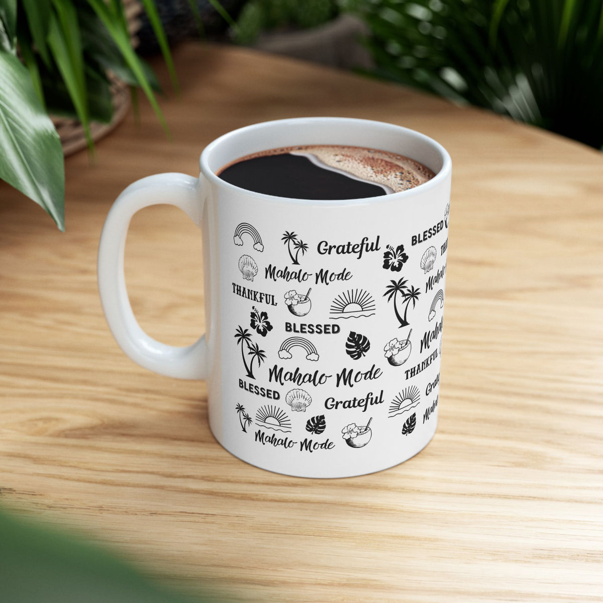 A white ceramic mug with a black tropical-themed print featuring the words 'Mahalo Mode', 'Grateful', and 'Blessed' alongside palm trees, monstera leaves, hibiscus flowers, and sun motifs, shown on a wood table with plants.