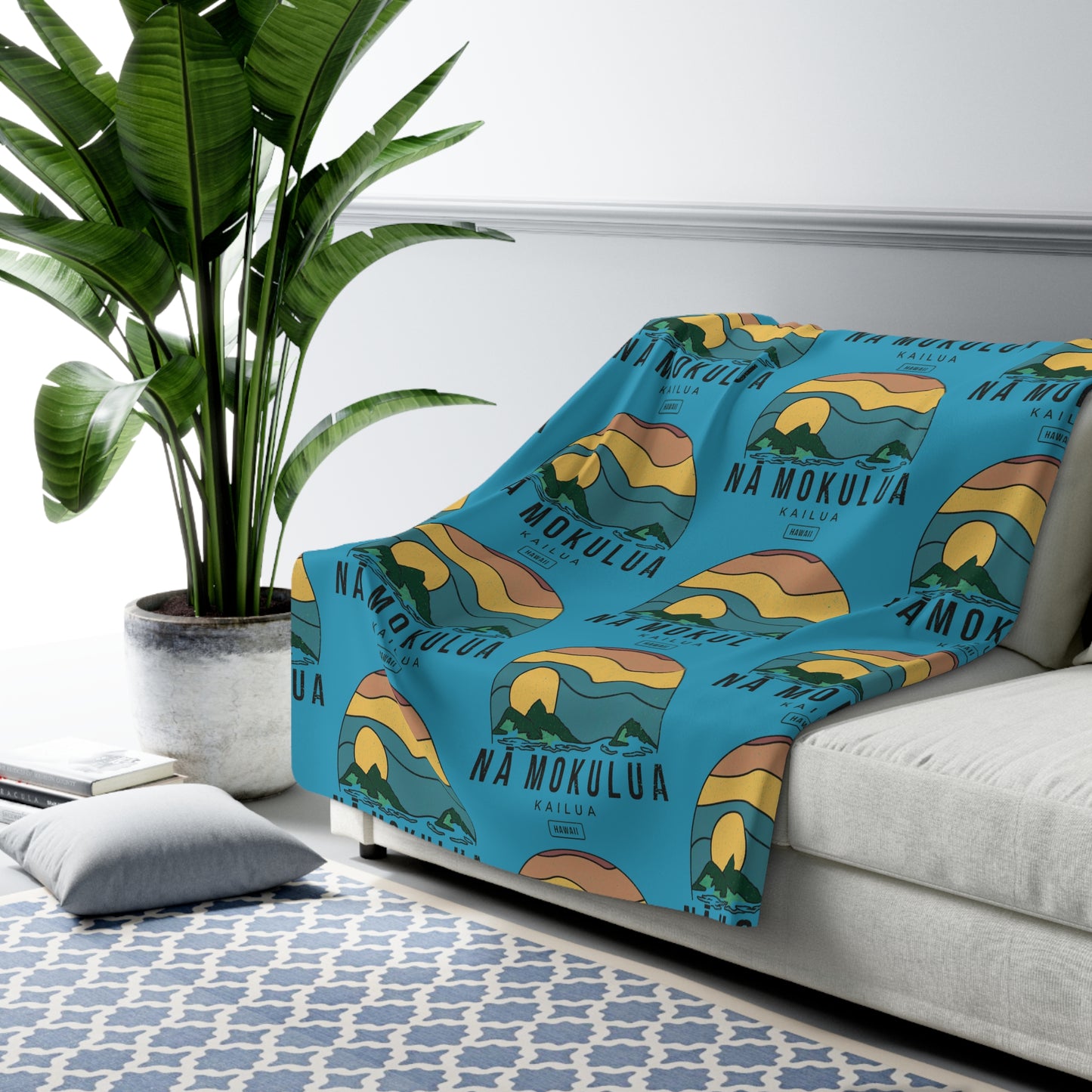 A blue throw blanket with a repeating 'Na Mokula' sunset landscape print is draped over a light gray sofa in a modern living room setting next to a large green potted plant.