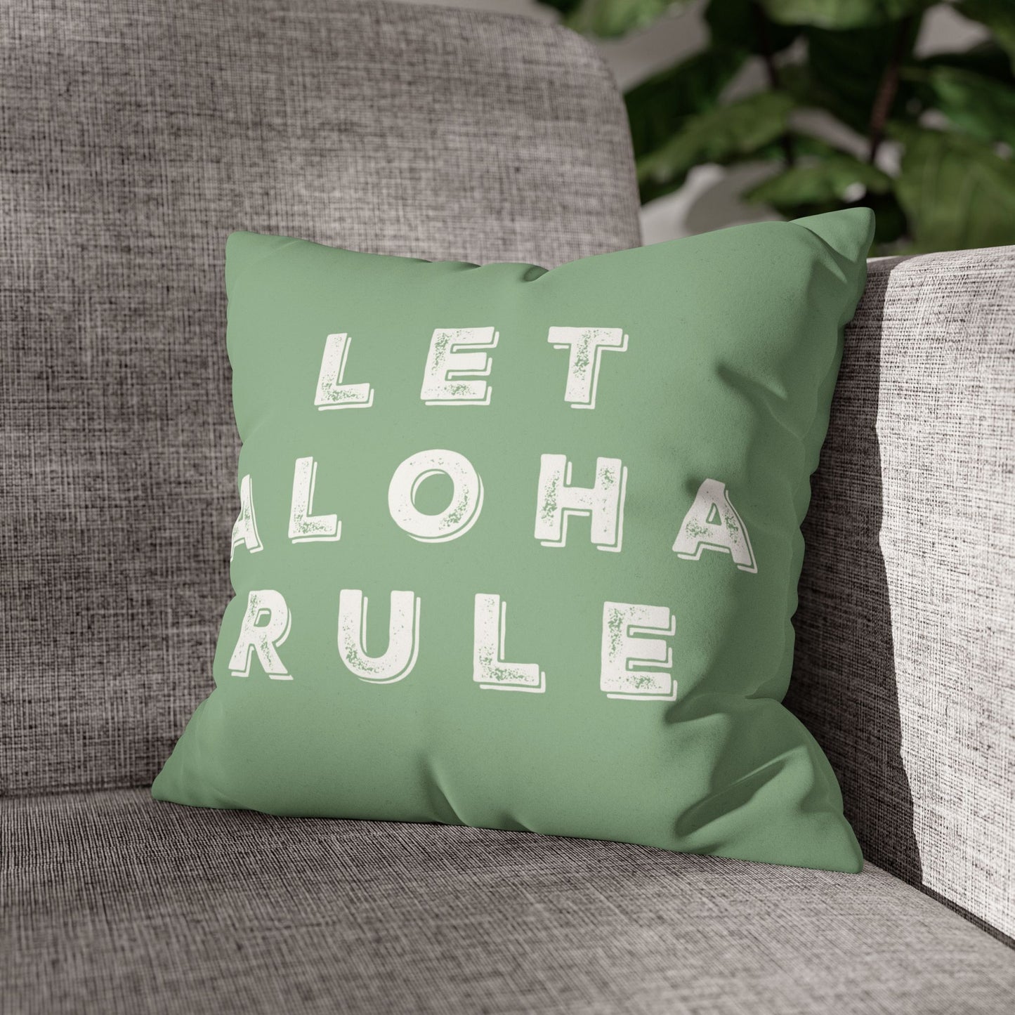 A square mint green throw pillow with the words 'LET ALOHA RULE' printed in distressed white block letters, resting on a gray textured armchair.