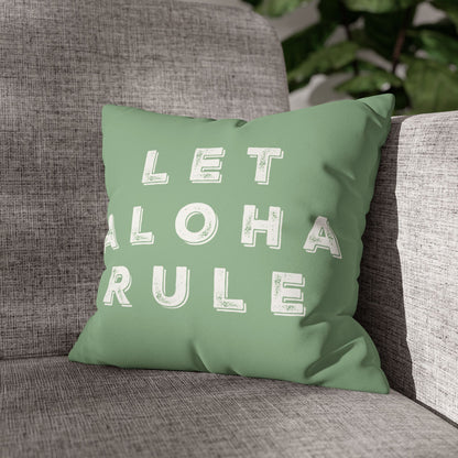 A square mint green throw pillow with the words 'LET ALOHA RULE' printed in distressed white block letters, resting on a gray textured armchair.