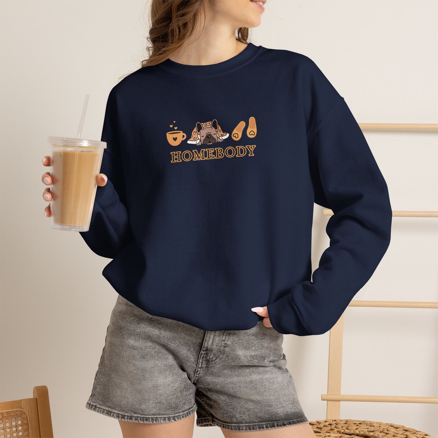 Person holding an iced coffee standing wearing a navy crewneck sweatshirt featuring a central graphic of a dog under a blanket, house slippers, coffee cups and the word 'HOMEBODY' printed below it.