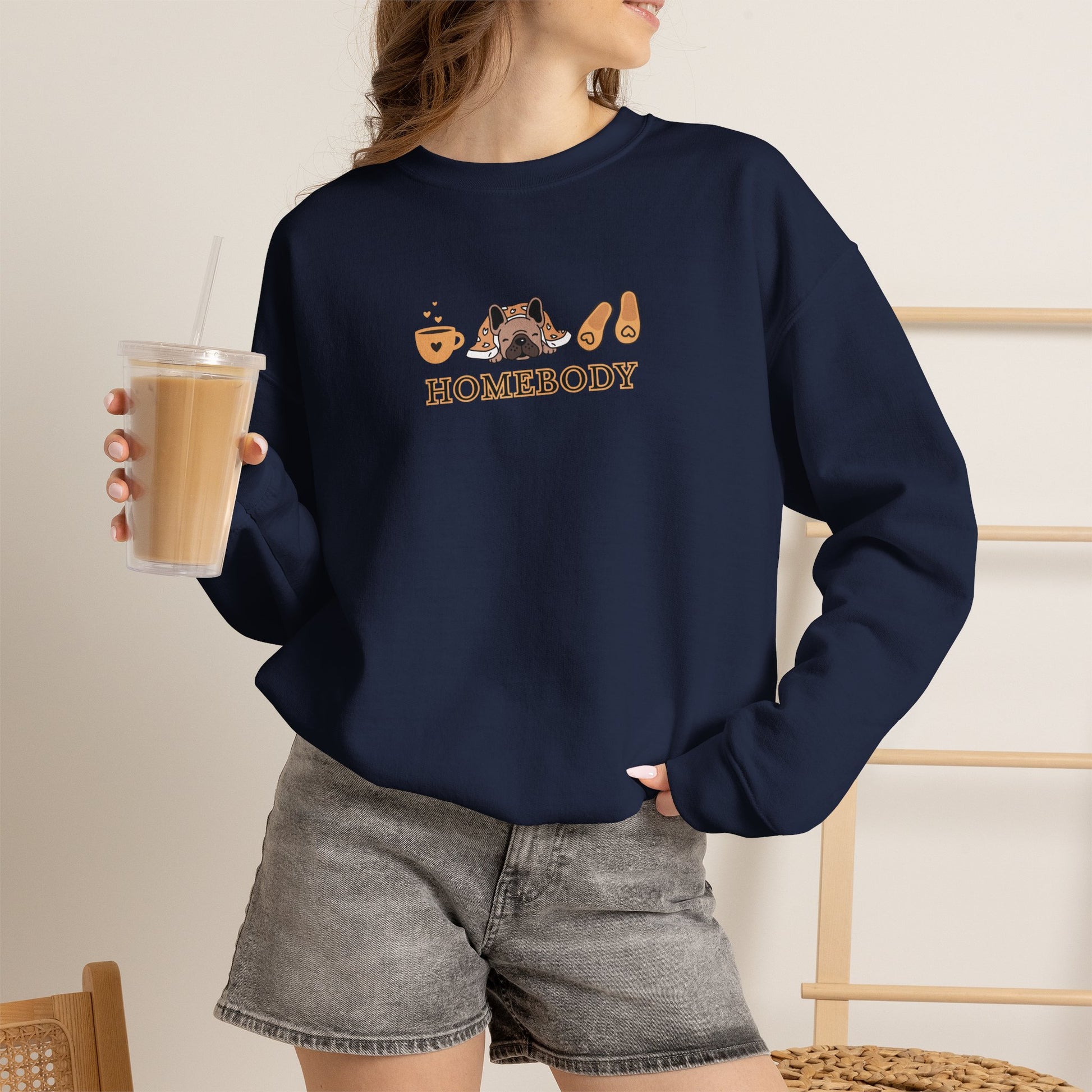 Person holding an iced coffee standing wearing a navy crewneck sweatshirt featuring a central graphic of a dog under a blanket, house slippers, coffee cups and the word 'HOMEBODY' printed below it.