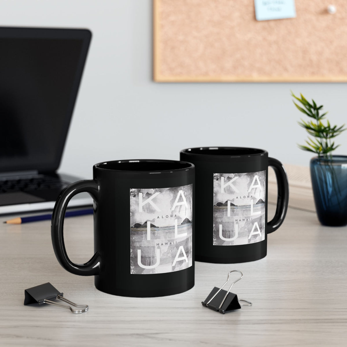 Two glossy black ceramic mugs featuring a square grayscale graphic print on the front with the words 'KAILUA', 'ALOHA', and 'HAWAII' over a landscape image, sitting on a desk with a computer and binder clips.