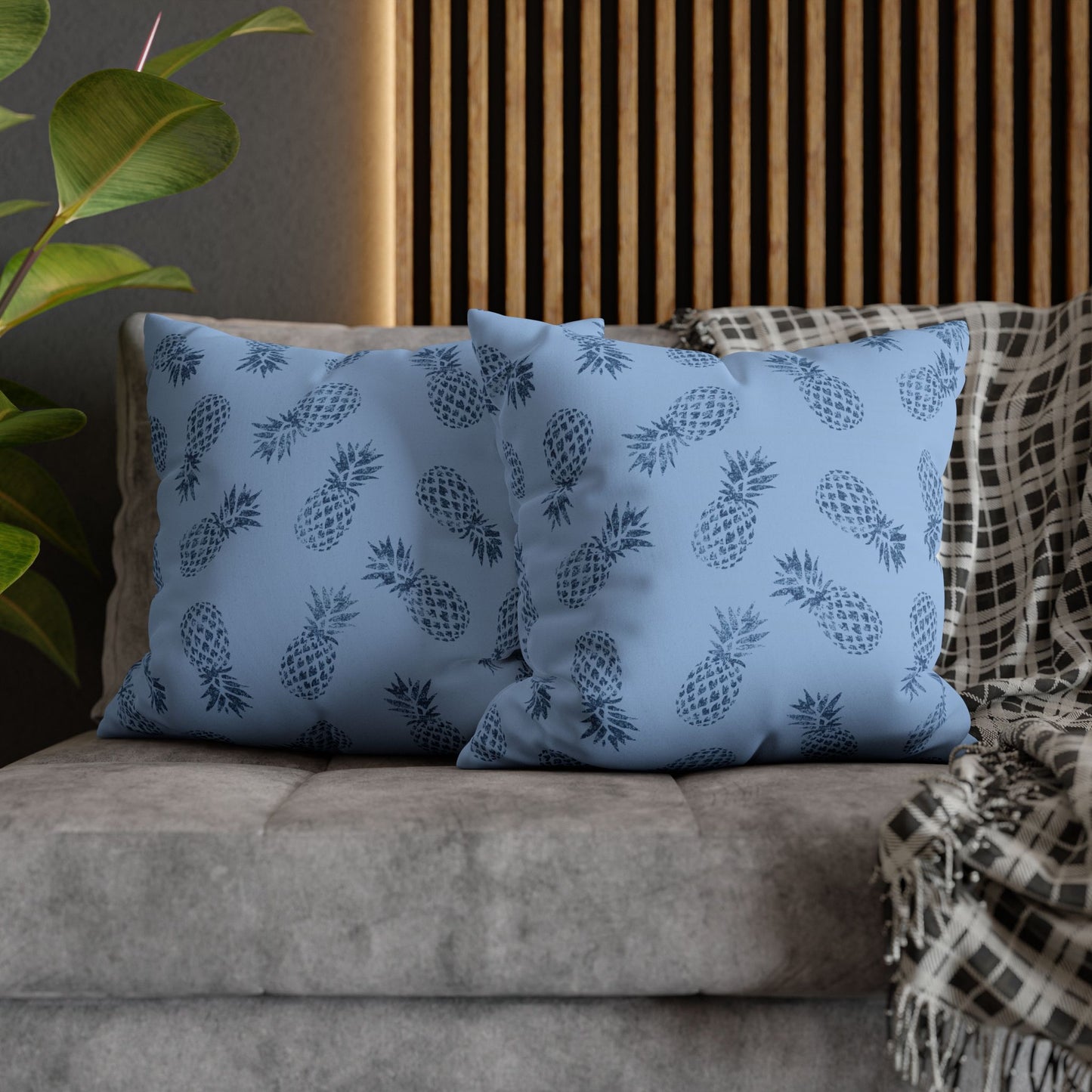 Two square throw pillows with a navy blue pineapple pattern on a dusty blue background, styled on a gray sofa with a plaid throw blanket and a wooden slat wall behind them.