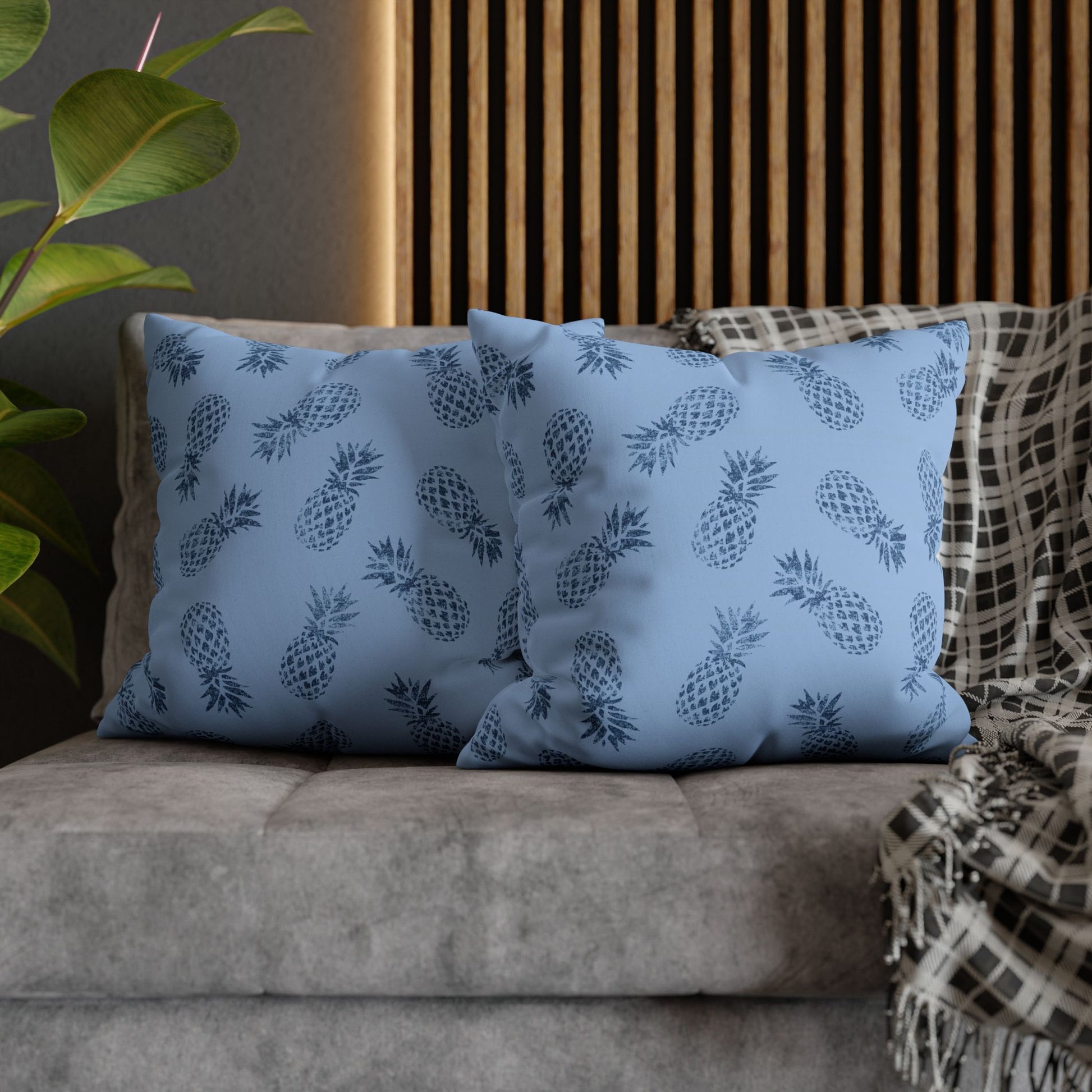 Two square throw pillows with a navy blue pineapple pattern on a dusty blue background, styled on a gray sofa with a plaid throw blanket and a wooden slat wall behind them.
