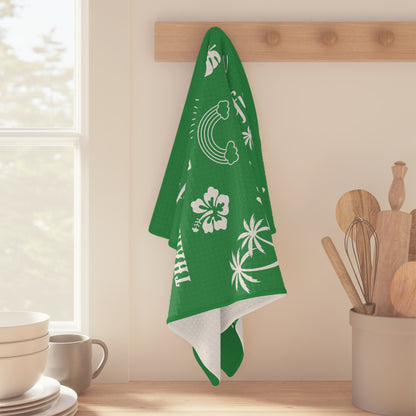 A green kitchen towel with white tropical and Hawaiian-themed prints hangs from a wooden rack on a kitchen wall. The towel features white palm trees, hibiscus flowers, a rainbow, and the word 'Hawaii'.