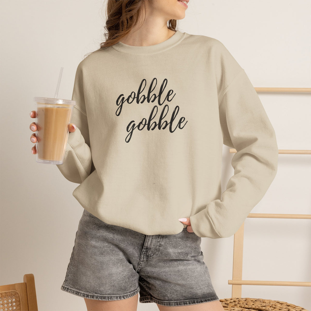 A woman wearing a beige crewneck sweatshirt with a graphic that reads 'Gobble Gobble' in black. She is holding a iced coffee and wearing gray denim shorts in a brightly lit indoor setting.